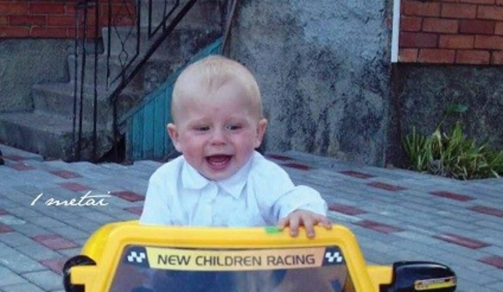 Smiling baby in a white shirt sits in a yellow toy car labeled 'NEW CHILDREN RACING' outdoors on a paved area with steps and brick wall behind him.