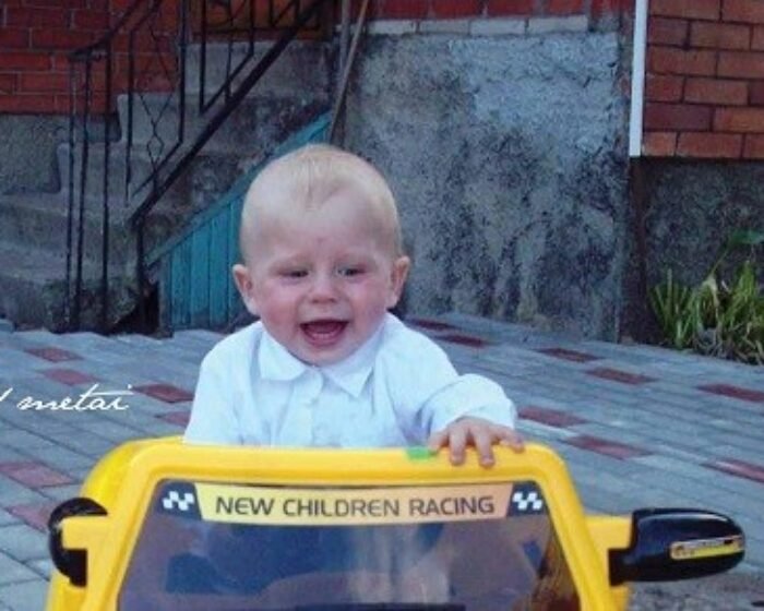 Smiling baby in a white shirt sits in a yellow toy car labeled 'NEW CHILDREN RACING' outdoors on a paved area with steps and brick wall behind him.