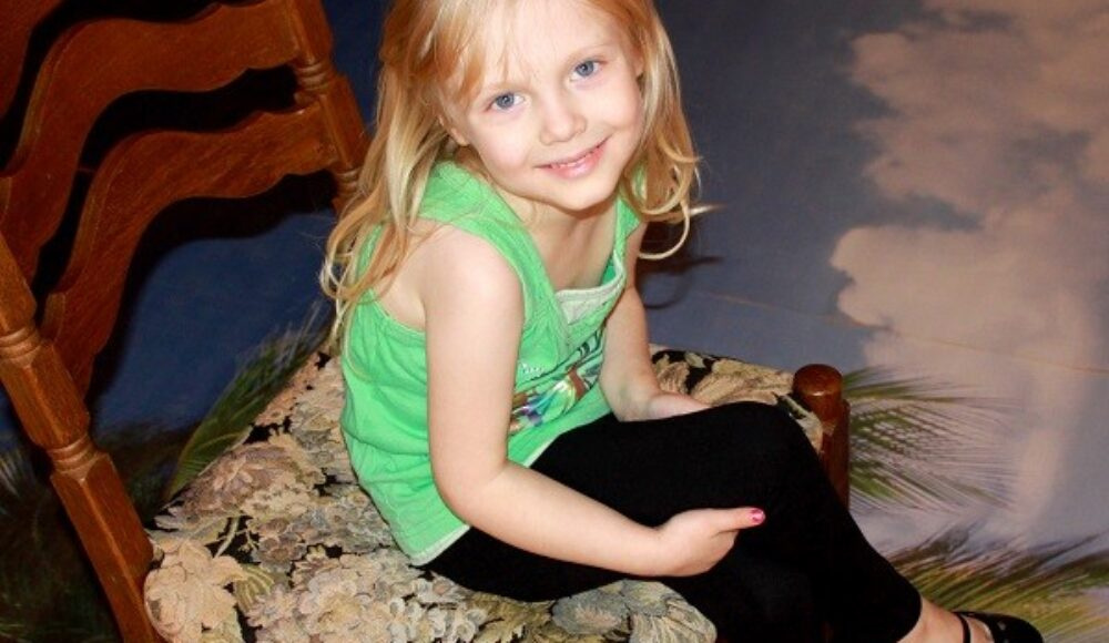 Smiling young girl with blonde hair sits sideways on a floral upholstered chair indoors, wearing a green top and black leggings.