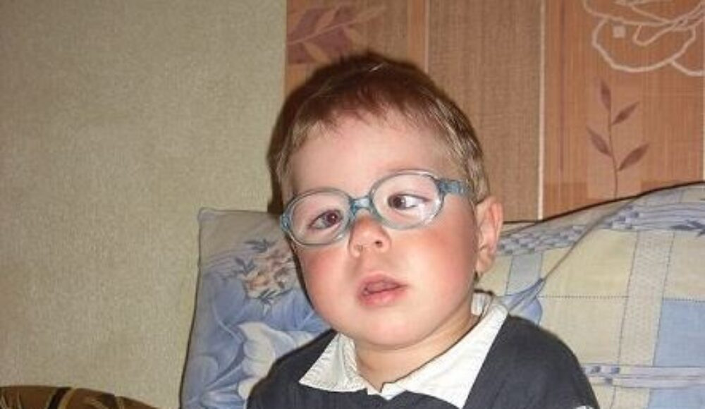 Young child with blue-framed glasses sits on a patterned sofa, looking toward the camera.