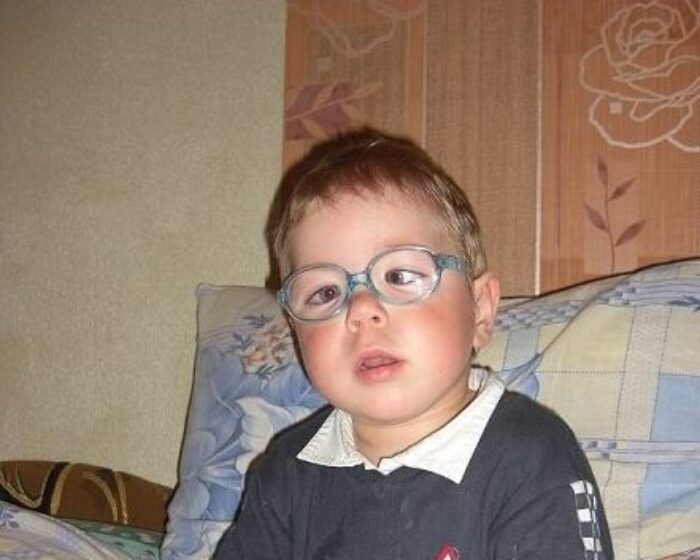 Young child with blue-framed glasses sits on a patterned sofa, looking toward the camera.