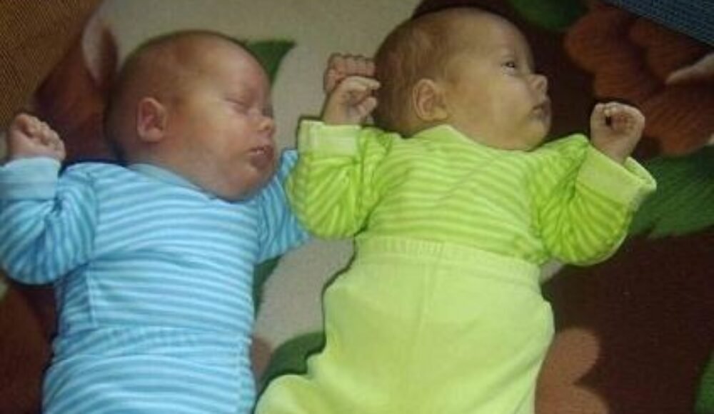 Two sleeping newborn twins lying side by side, one in a blue striped onesie and the other in a lime-green striped onesie.