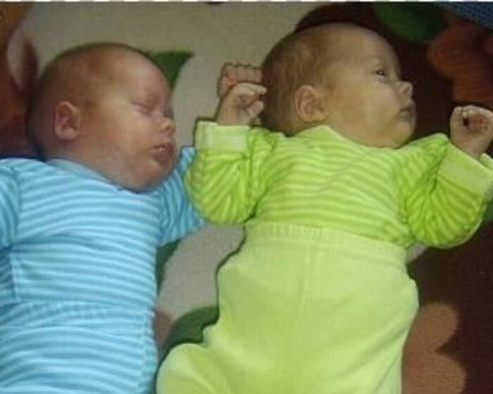 Two sleeping newborn twins lying side by side, one in a blue striped onesie and the other in a lime-green striped onesie.