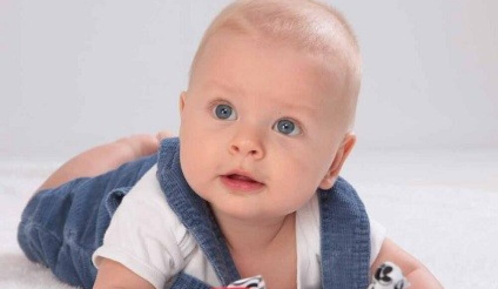 Baby lying on tummy on a white surface, wearing a white onesie and blue overalls, looking up with blue eyes while playing with a colorful toy.