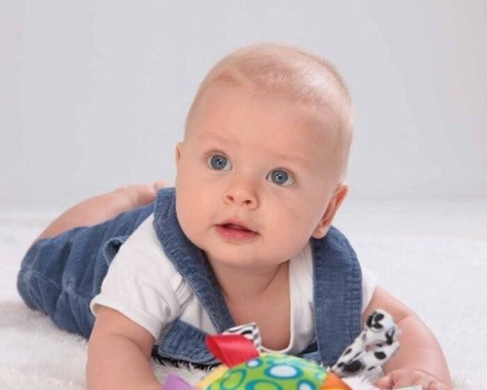 Baby lying on tummy on a white surface, wearing a white onesie and blue overalls, looking up with blue eyes while playing with a colorful toy.