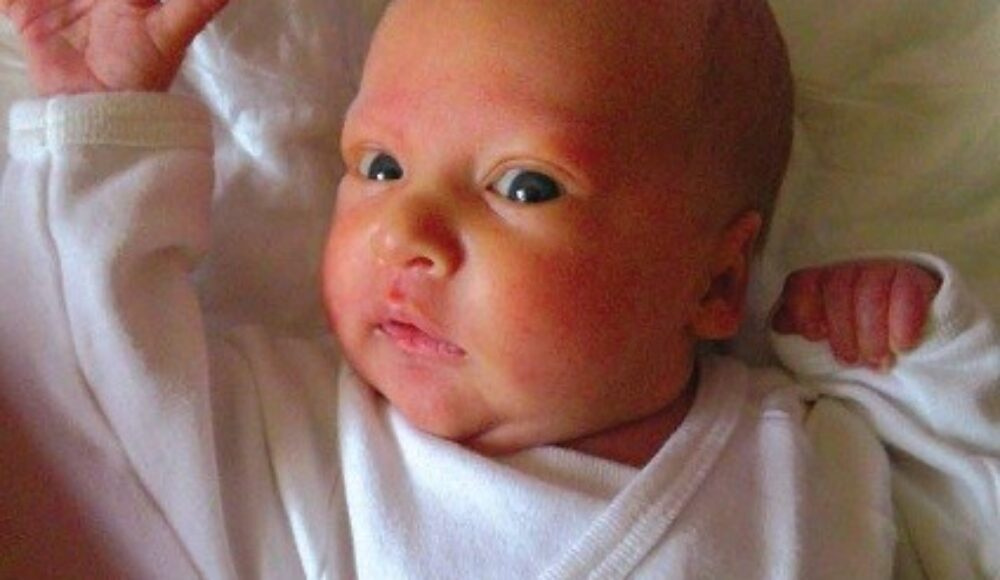 Close-up of a newborn baby lying on a pale bed, wearing a white outfit, with arms raised and a curious expression.