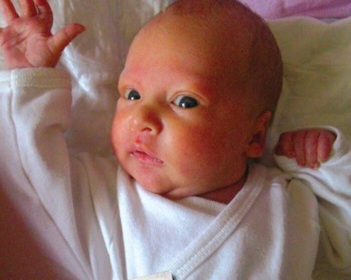 Close-up of a newborn baby lying on a pale bed, wearing a white outfit, with arms raised and a curious expression.