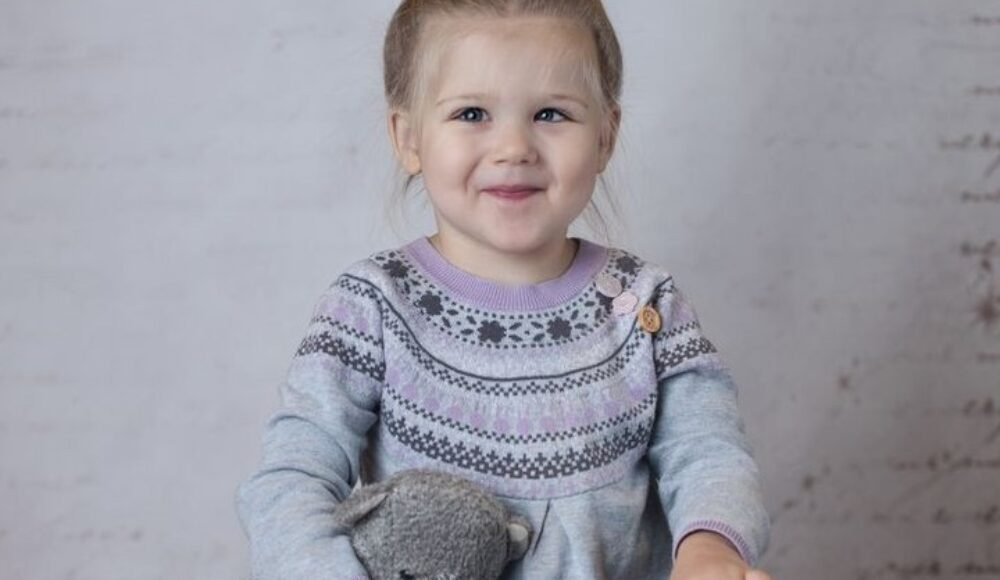 Young girl with a gray sweater holding a stuffed animal and a framed photo, posing in front of a light backdrop.
