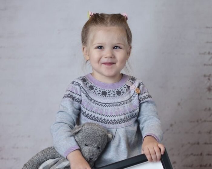 Young girl with a gray sweater holding a stuffed animal and a framed photo, posing in front of a light backdrop.