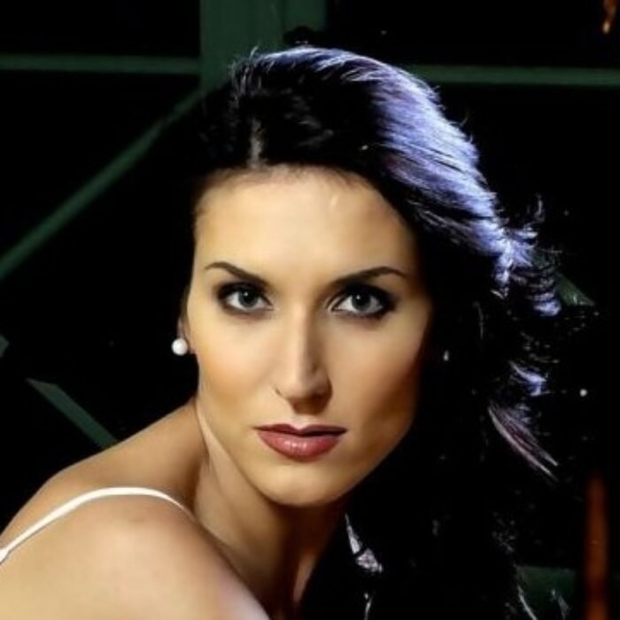 Close-up portrait of a woman with dark hair, pearl earrings, and striking makeup looking at the camera.