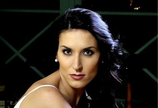 Close-up portrait of a woman with dark hair, pearl earrings, and striking makeup looking at the camera.