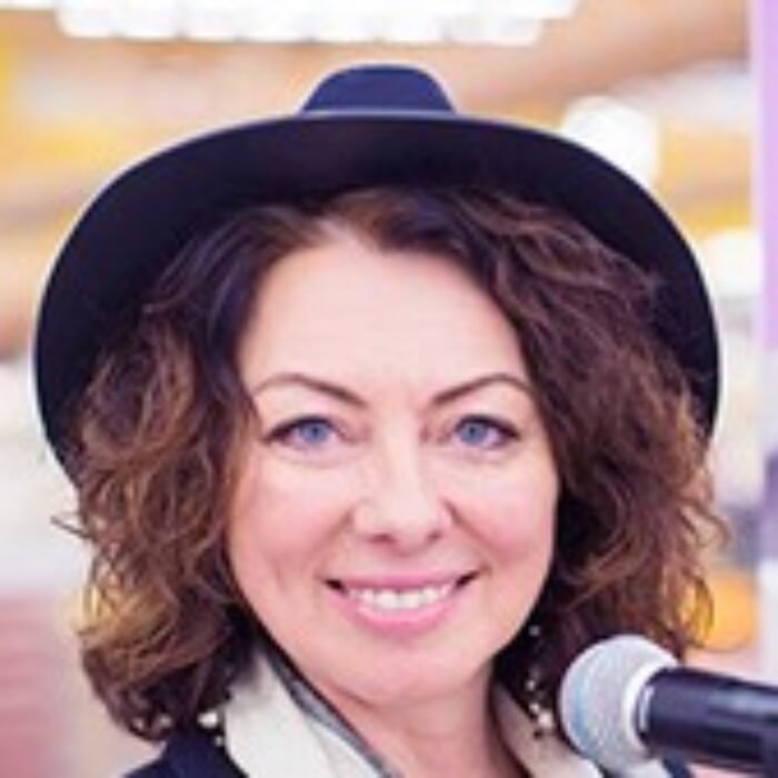 Smiling woman wearing a dark hat and scarf speaks into a microphone at a public event.