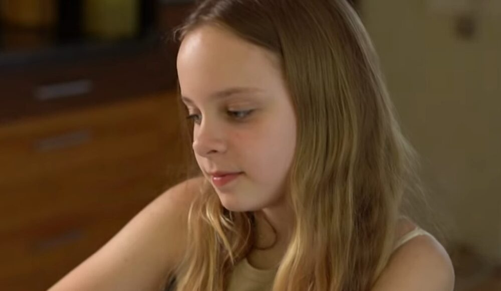 Young girl with long blond hair looking downward indoors, wearing a light tank top.