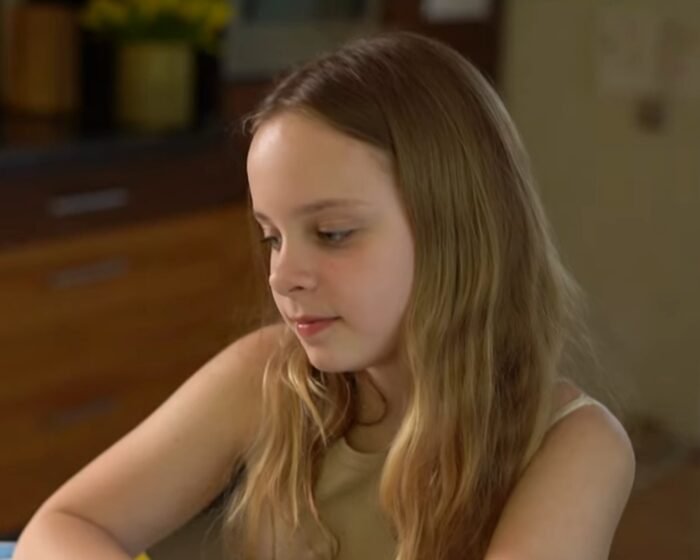 Young girl with long blond hair looking downward indoors, wearing a light tank top.