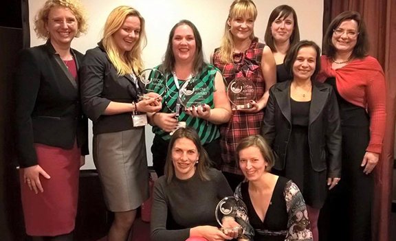 Nine women in formal attire posing with glass awards at an awards ceremony.