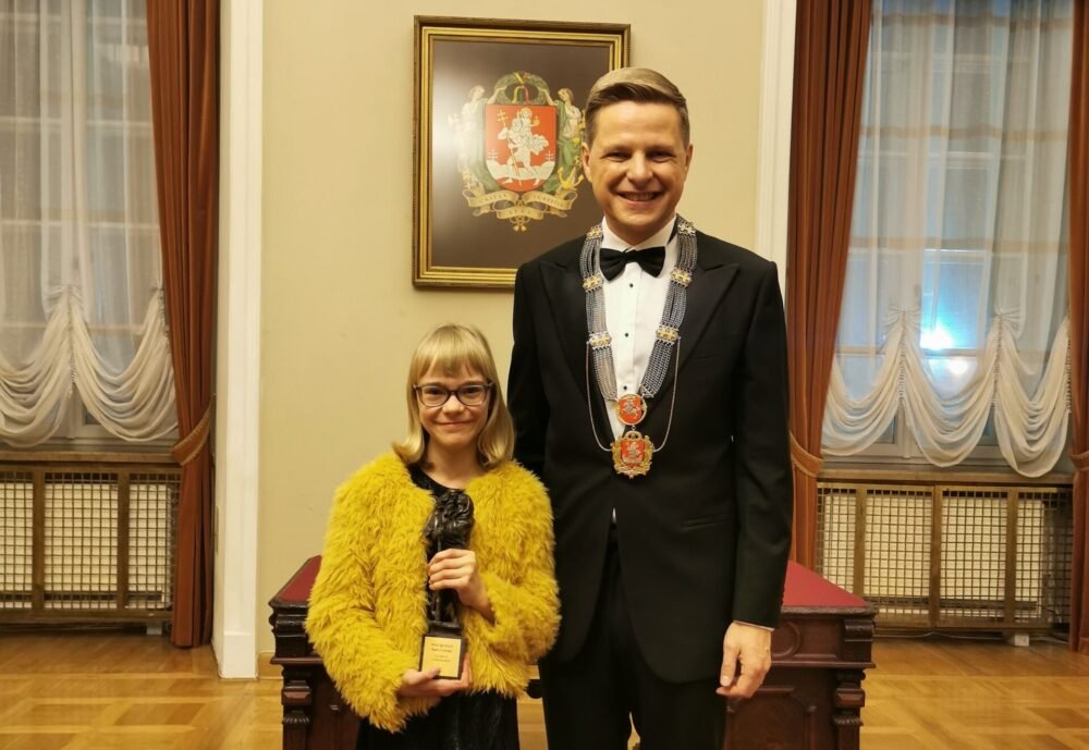 Young girl in a yellow fur jacket holds a trophy while standing next to a man in a black tuxedo wearing a ceremonial chain, in a formal room with draped curtains and a crest on the wall.