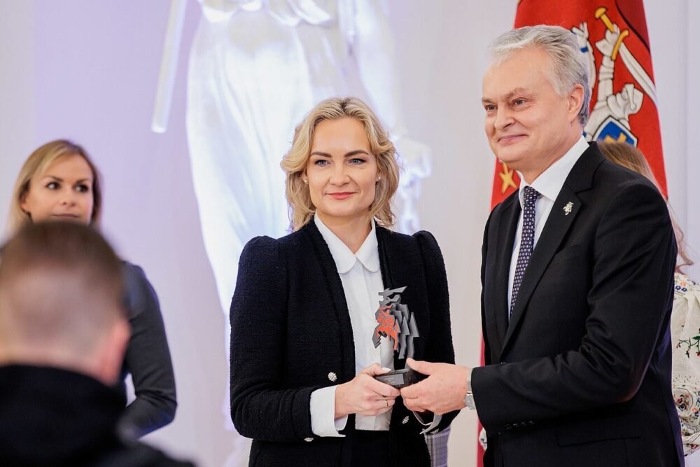 Woman in a black blazer accepts a trophy from a smiling man in a suit at a formal award ceremony, with a flag in the background.