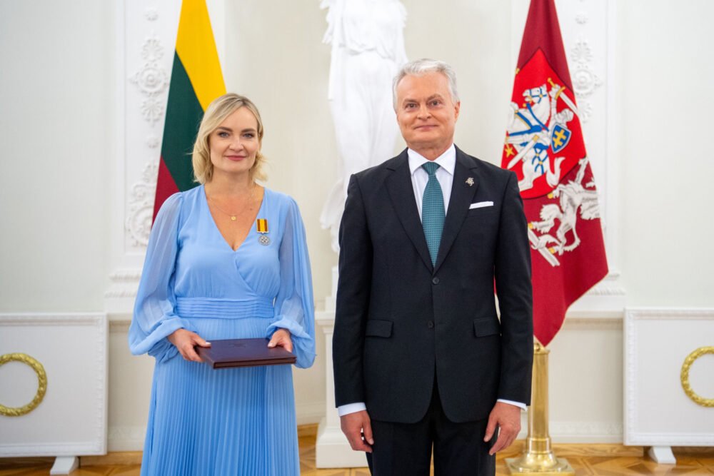 Formal portrait of a blonde woman in a light blue gown holding a brown folder/award, standing beside a man in a dark suit, with flags in the background.