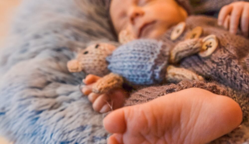 Sleeping baby wrapped in a knitted brown blanket, resting in a wicker basket with plush toys nearby