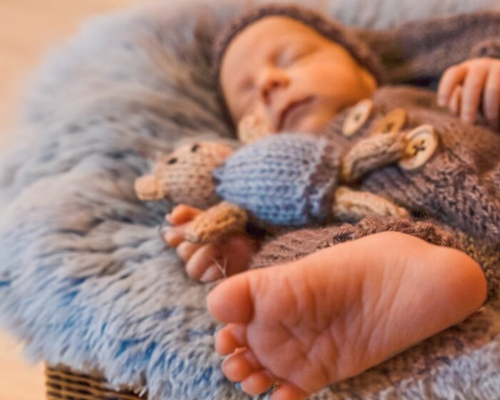 Sleeping baby wrapped in a knitted brown blanket, resting in a wicker basket with plush toys nearby