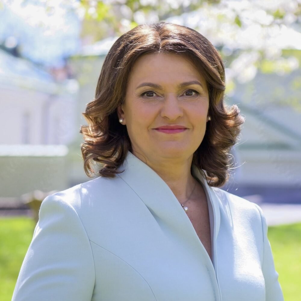 Portrait of a smiling woman outdoors in a light blue coat with green, sunlit background blur behind her