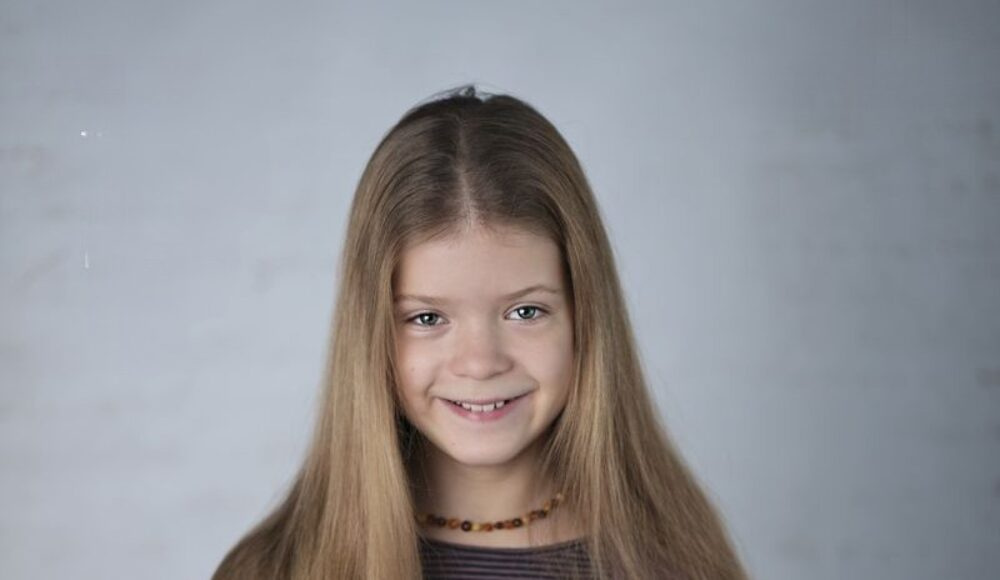 Portrait of a smiling girl with long light brown hair against a neutral gray background, wearing a striped shirt and a beaded necklace.