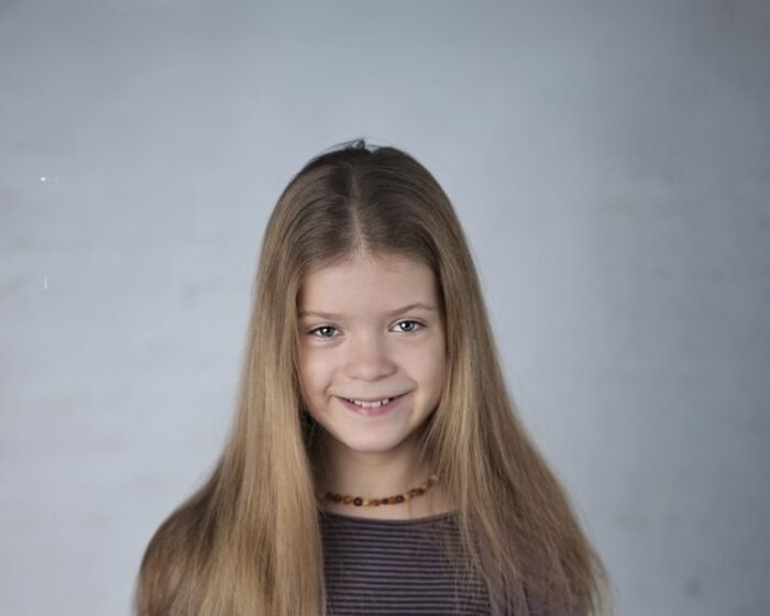 Portrait of a smiling girl with long light brown hair against a neutral gray background, wearing a striped shirt and a beaded necklace.
