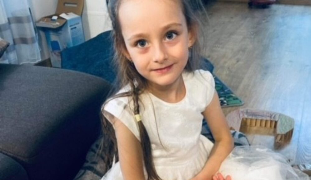 Young girl in a white dress sitting on the floor at home, facing the camera.