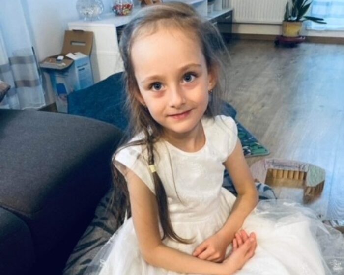 Young girl in a white dress sitting on the floor at home, facing the camera.
