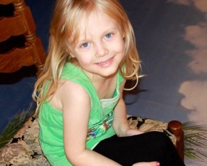 Smiling blonde girl in a green tank top sits on a patterned chair indoors.