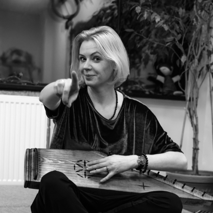 Woman with short blonde hair sits cross‑legged, pointing at the camera while holding a wooden stringed instrument on her lap.