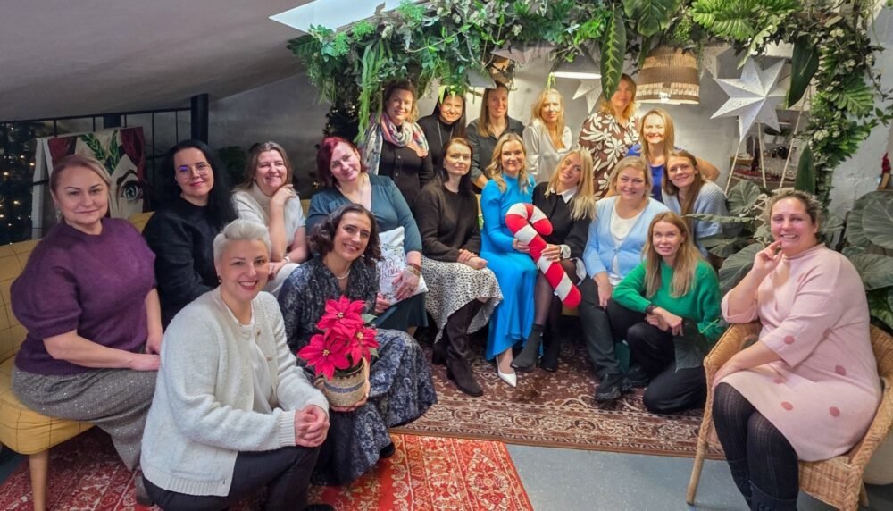 Group of women posing together for a festive indoor photo, surrounded by greenery and holiday decor.