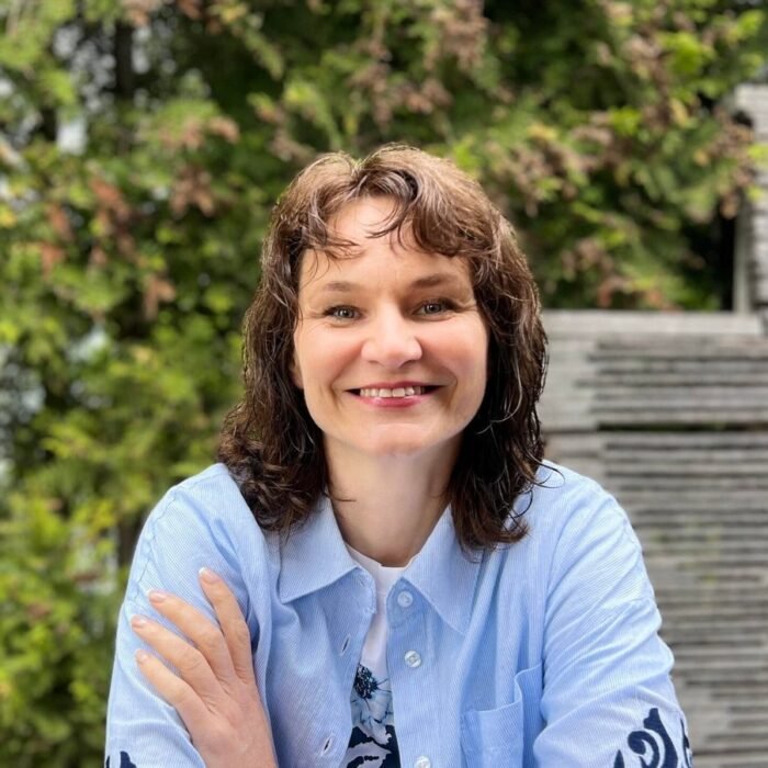 Smiling woman with shoulder-length brown hair in a light blue button-down shirt, outdoors with greenery and wooden steps behind her.