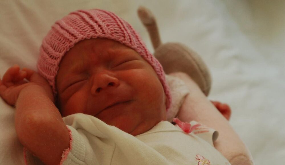 Newborn baby wearing a pink knit hat, resting with arms raised on a light blanket.