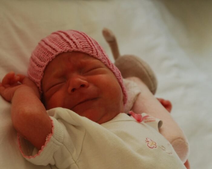 Newborn baby wearing a pink knit hat, resting with arms raised on a light blanket.