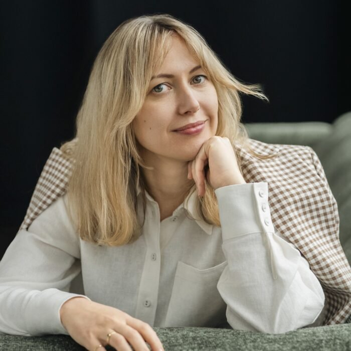 Woman with shoulder-length blonde hair resting her chin on her hand, wearing a white shirt with a brown-and-white checkered jacket draped over her shoulders, seated on a sofa.