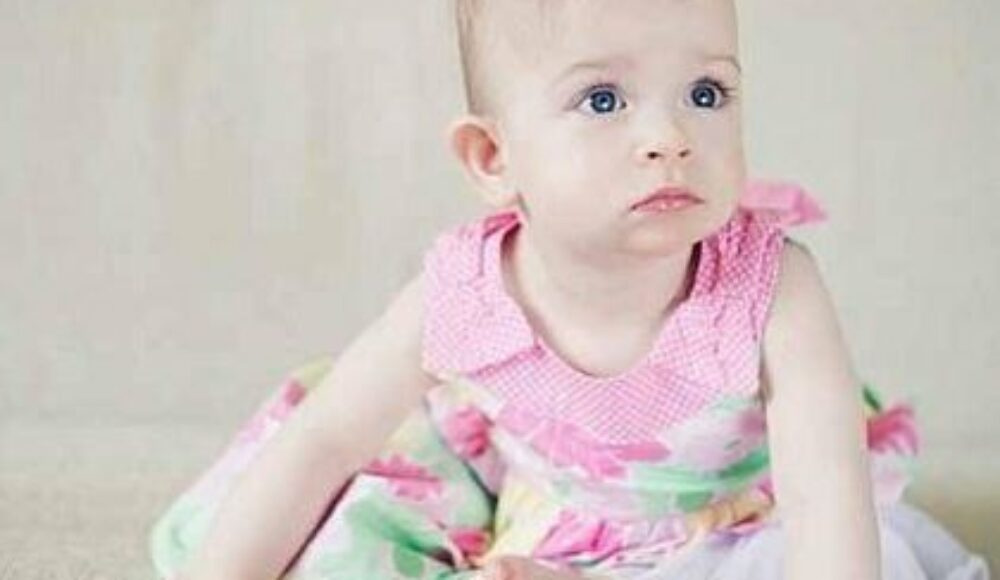 Baby girl in a pink floral dress with a bow in her hair, sitting on a light carpet and gazing to the side?