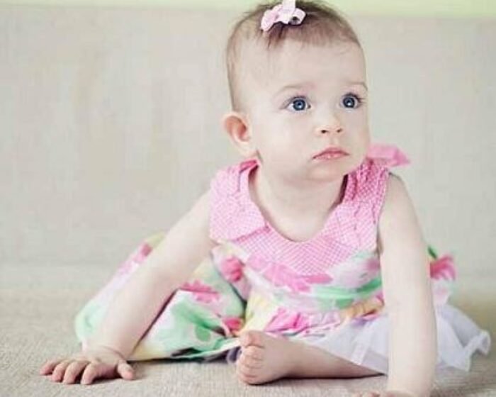 Baby girl in a pink floral dress with a bow in her hair, sitting on a light carpet and gazing to the side?