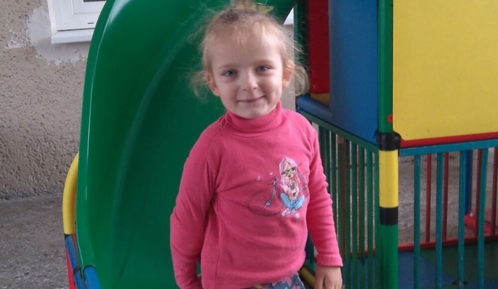 Young girl in a pink sweater stands at a colorful playground slide, smiling at the camera.