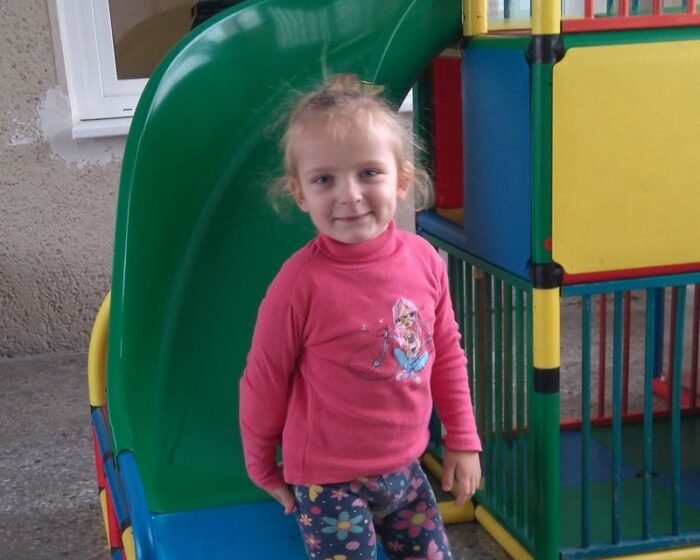 Young girl in a pink sweater stands at a colorful playground slide, smiling at the camera.