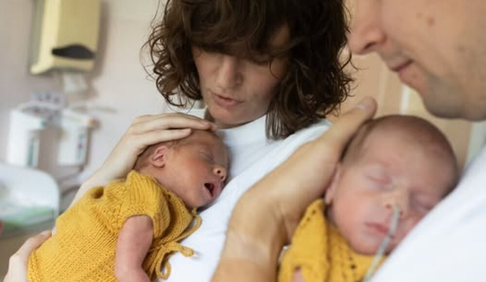 Mother gently cradles a newborn twin in a mustard outfit while the father holds the other baby in a hospital room, both babies dressed in mustard.