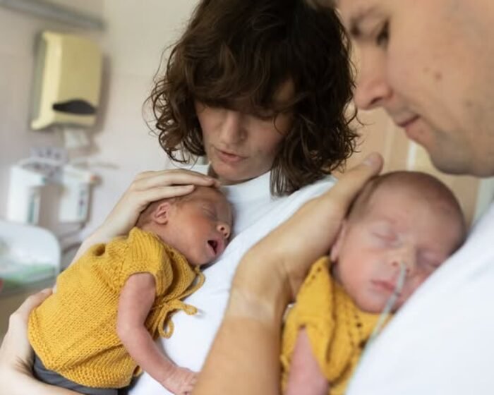 Mother gently cradles a newborn twin in a mustard outfit while the father holds the other baby in a hospital room, both babies dressed in mustard.