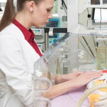 Young healthcare worker in a white coat and red shirt tending a newborn in a clear incubator, with medical monitors in the background.