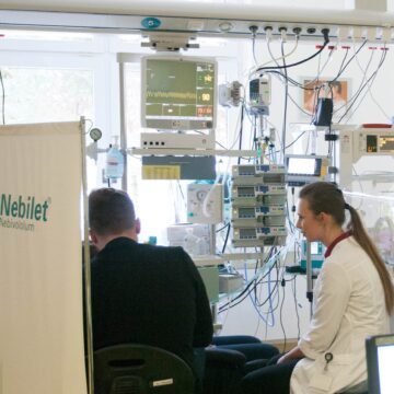 Medical staff seated at a patient monitoring station in an ICU with multiple monitors, IV pumps, and tubing surrounding them