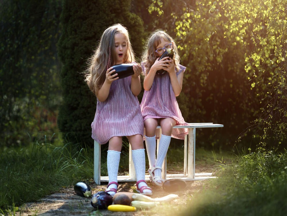 Two young girls in pink striped dresses sit on a wooden bench outdoors, playing with colorful plastic toys on the ground.