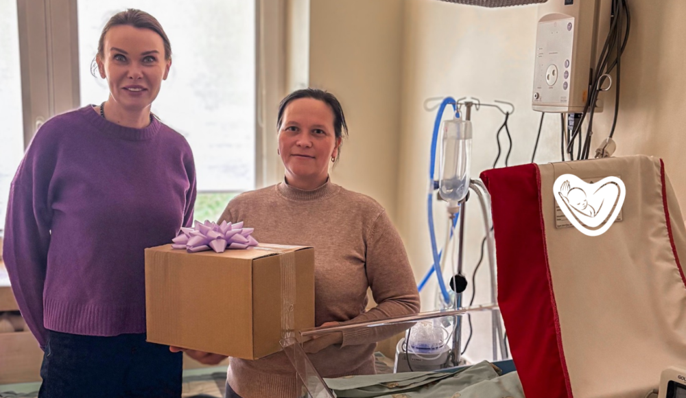 Two women in a hospital room, one handing a gift box with a purple bow to the other near a hospital bed and IV stand.