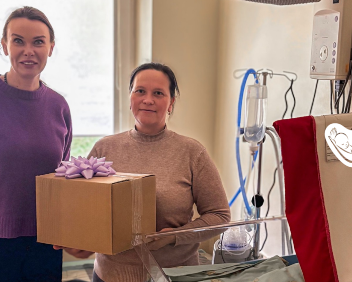 Two women in a hospital room, one handing a gift box with a purple bow to the other near a hospital bed and IV stand.