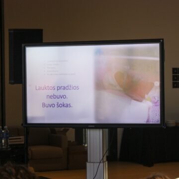 Large presentation screen in a dim conference room showing a slide with Lithuanian text; a laptop and water bottles sit on a table to the left.