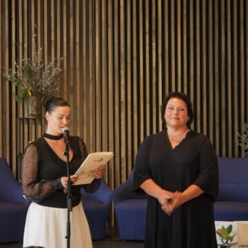 Two women on a stage: one reads from a booklet into a microphone while the other smiles, with a brown slatted wall behind them.