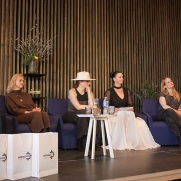 Panel of six women seated on blue chairs on a stage, with gift bags and potted plants in front and a dark wooden slat backdrop.